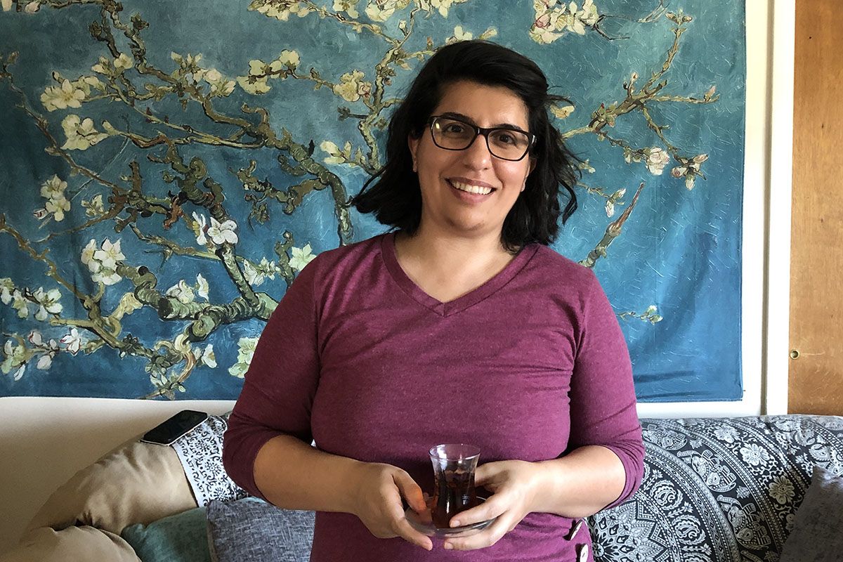 Deyra Dogan standing in front of a blue floral wall hanging holding a glass cup of tea
